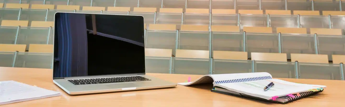 Open laptop, notebooks, and papers on a desk in an empty lecture hall with rows of empty seats in the background.