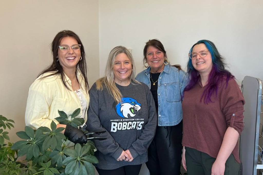 Four women are standing indoors, smiling at the camera, with a green plant in the foreground.