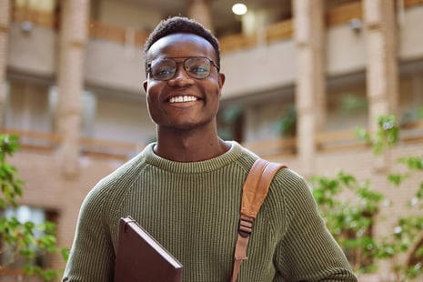 A young man wearing glasses and a green sweater smiles while holding a laptop and carrying a brown backpack, possibly preparing for his online human services associate degree, standing in a bright indoor space with brick walls and plants.