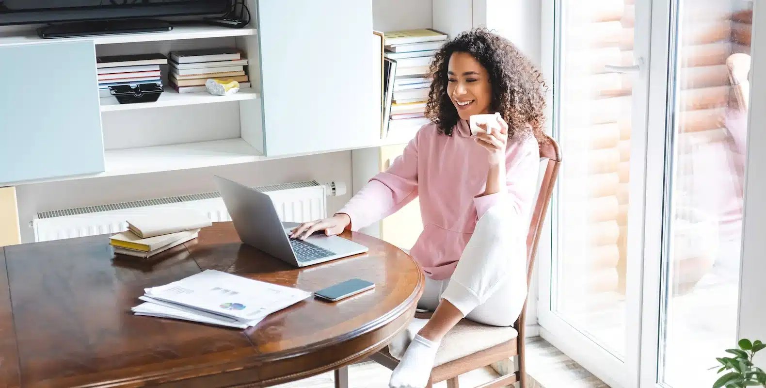 Woman in pink hoodie sitting at a table with a laptop, holding a coffee cup, and smiling in a bright room.