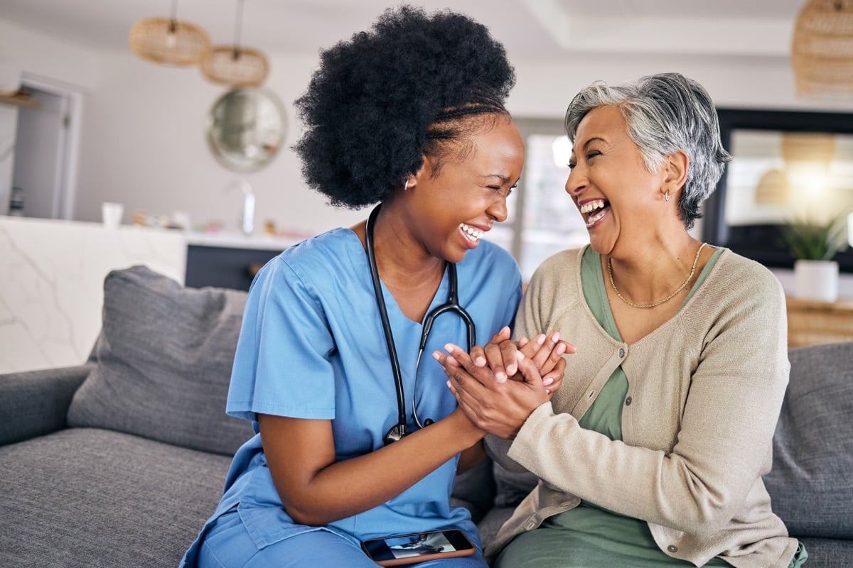 A nurse and an elderly woman sitting on a couch, holding hands and laughing together in a cozy, bright living room.