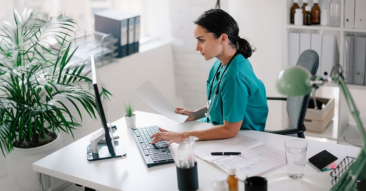 A healthcare professional in scrubs works on a computer while holding documents in a medical office.