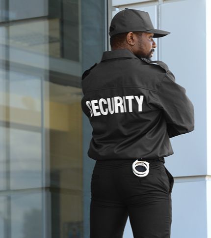A Security Guard in a black uniform and hat stands outside a modern building. The word "SECURITY" is printed on the back of his jacket, and handcuffs are attached to his belt.