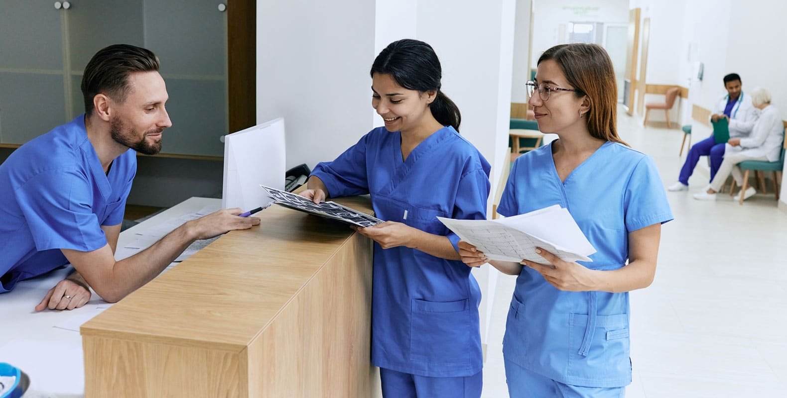 Three nurses in blue scrubs consult documents at a hospital desk, representing the duty of care in nursing and teamwork in healthcare environments.