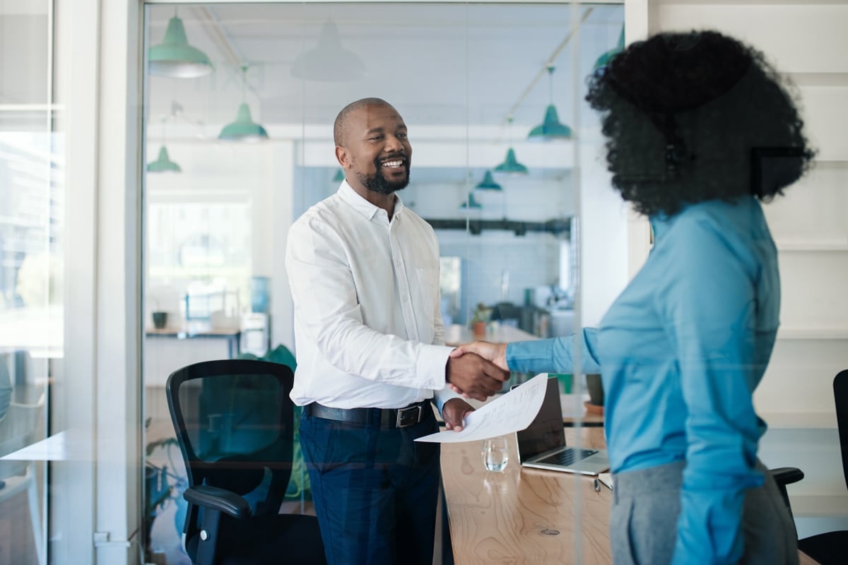 Two people shaking hands in an office setting, both smiling, one holding documents.