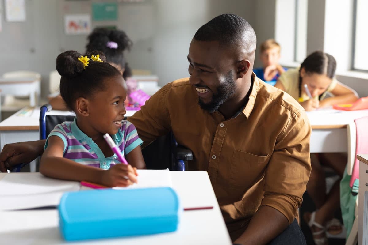 A smiling man with an Early Childhood Certificate sits beside a young girl in a classroom, both engaged in conversation, surrounded by other children.