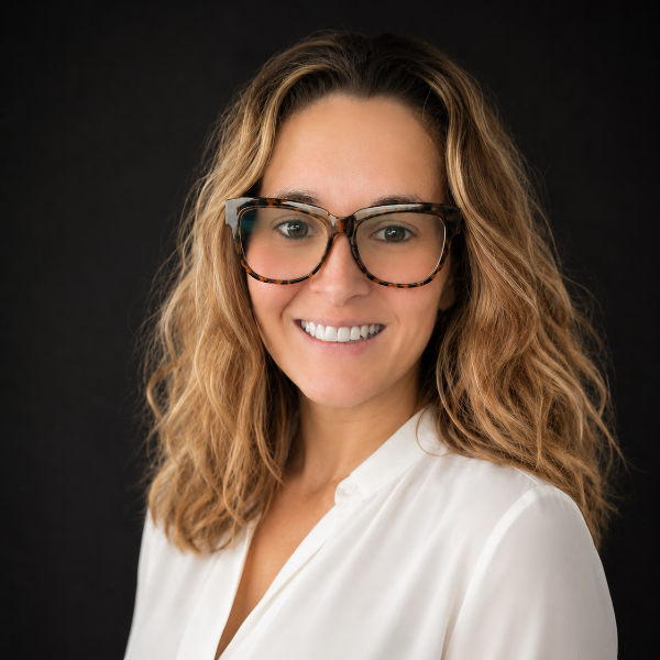 A woman with wavy hair and tortoiseshell glasses smiles at the camera against a dark background.