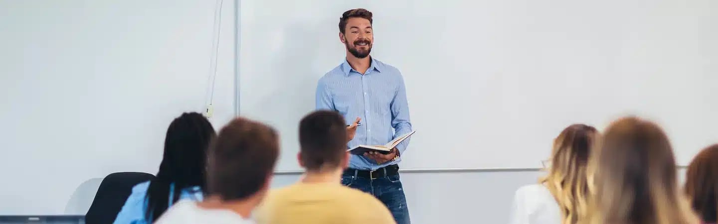 A man stands at the front of a classroom holding a book, smiling and speaking to a group of seated students.