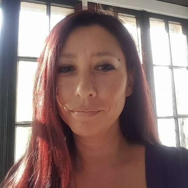 A woman with long, straight reddish-brown hair is smiling softly. She is indoors, standing in front of tall windows with sunlight streaming in behind her.