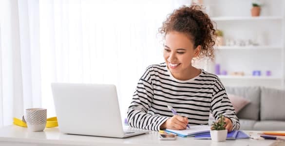 Woman smiling, taking notes while looking at a laptop in a bright room with coffee and potted plant on desk.