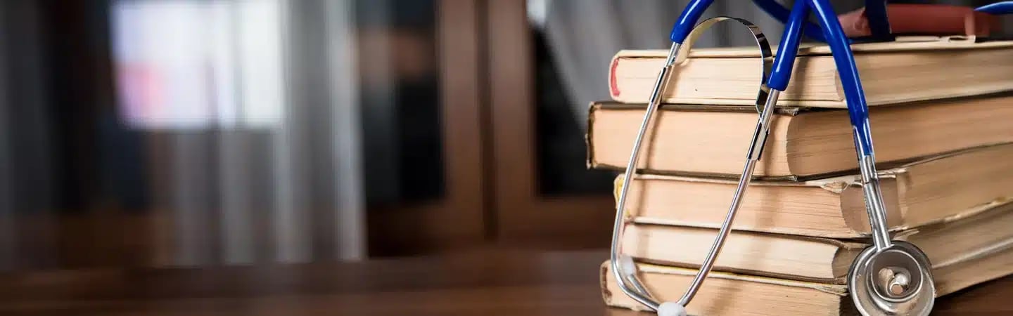 Stack of books on a wooden desk with a stethoscope draped over them, in front of a blurred background of windows and curtains.