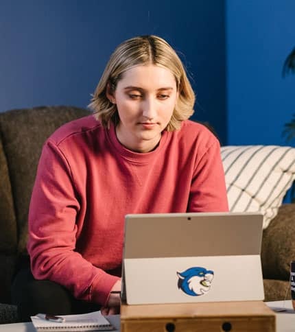 Person with short blonde hair wearing a red sweatshirt sits on a couch using a laptop. The laptop has a sticker of a cat face on it. There is a notebook and pen beside them, and a striped pillow is visible in the background.
