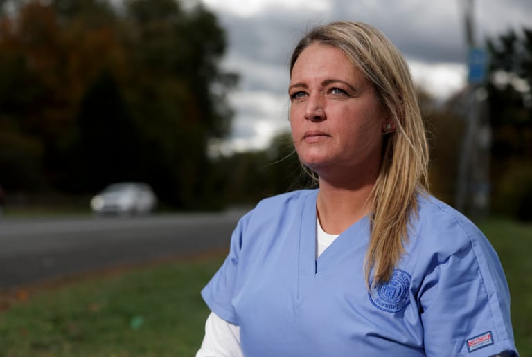 A woman in light blue medical scrubs stands outdoors near a road, looking thoughtfully into the distance. Trees and a cloudy sky are visible in the background, reflecting the spirit of Bryant & Stratton College’s commitment to online enrollment.