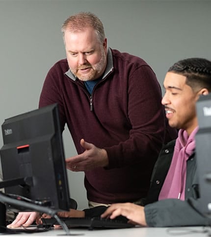 A bearded man in a maroon sweater assists a younger man with braided hair at a computer. Both are focused on the screen in a classroom or office setting.
