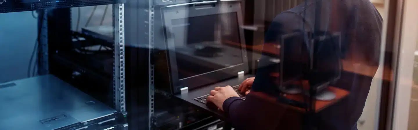 Person working on a laptop in a server room, surrounded by racks of network equipment.