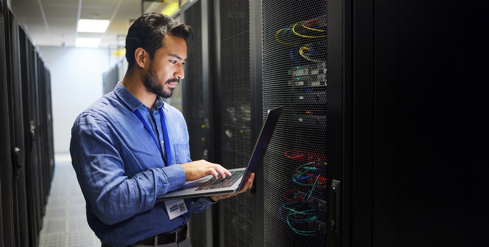 A person in a blue shirt stands in a server room, holding a laptop and looking at a server rack with cables and equipment. The surroundings are dimly lit with fluorescent lights above.
