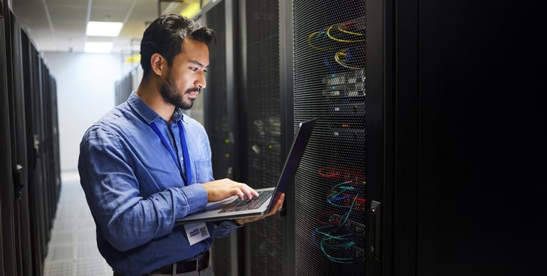 A person in a blue shirt stands in a server room, holding a laptop and looking at a server rack with cables and equipment. The surroundings are dimly lit with fluorescent lights above.