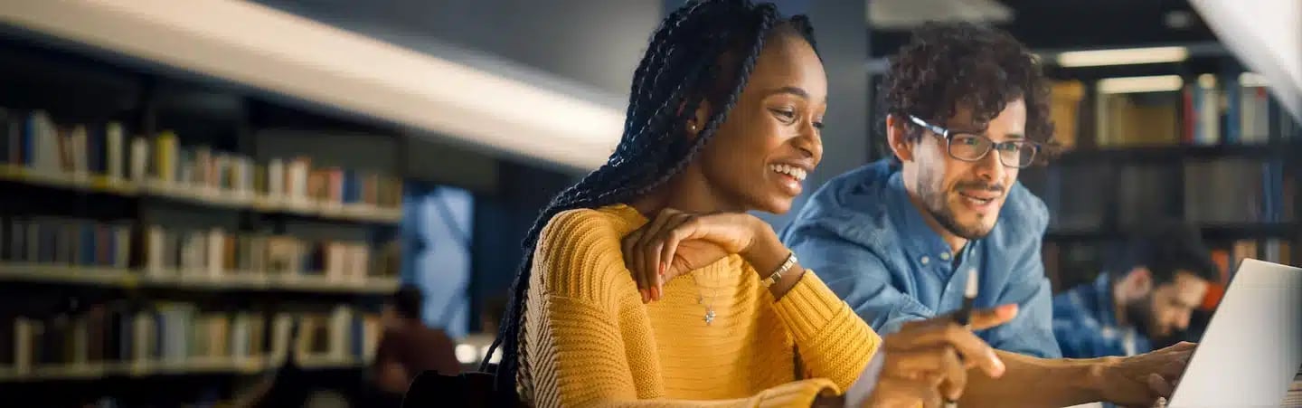 Two people sit at a table in a library, smiling and looking at a laptop, with bookshelves in the background.