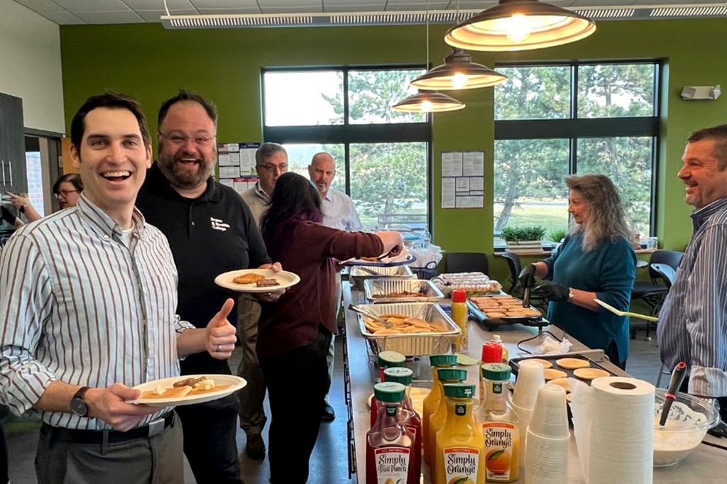 A group of people are smiling and serving food at a buffet in a well-lit room with large windows.