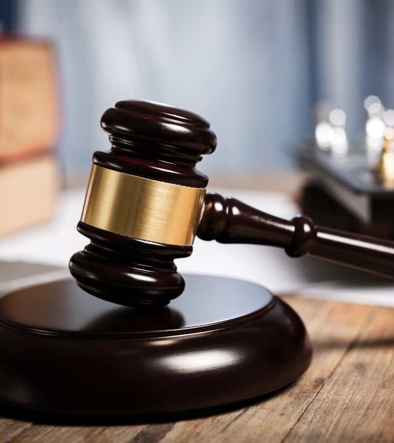 A wooden judge’s gavel with a brass band rests on a sound block on a wooden table, often used by a Sheriff, with blurred legal books and documents in the background.