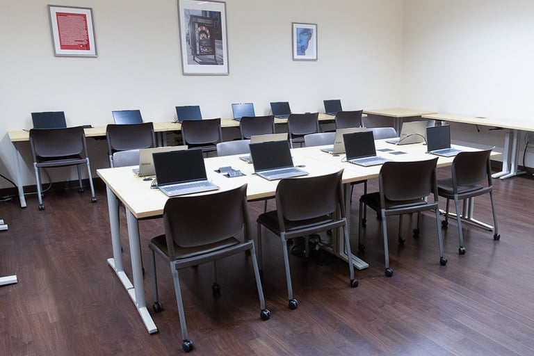 A classroom with wooden floors, several tables arranged in rows, brown chairs, and laptops on each table. Three framed pictures hang on the cream-colored walls.