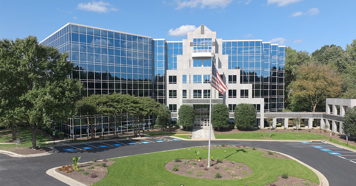 Modern office building with glass windows, surrounded by trees and landscaping, featuring a circular driveway and an American flag—perfect for institutions like Richmond College or colleges in Richmond VA. The sky is blue with scattered clouds.