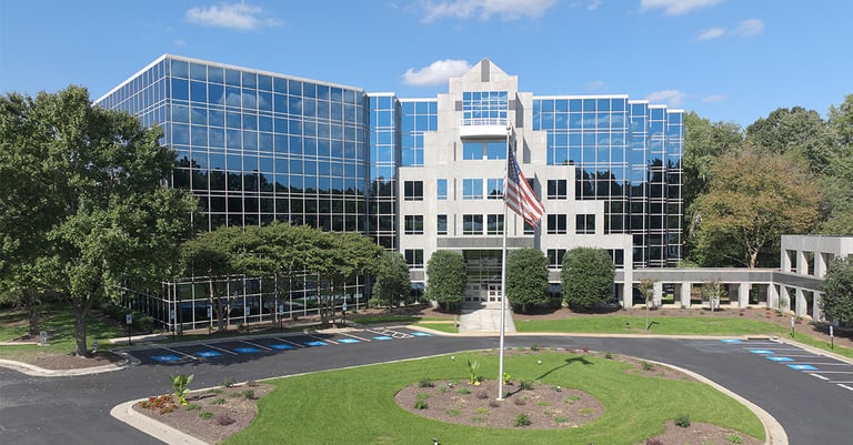 Modern office building with glass windows, surrounded by trees and landscaping, featuring a circular driveway and an American flag—perfect for institutions like Richmond College or colleges in Richmond VA. The sky is blue with scattered clouds.