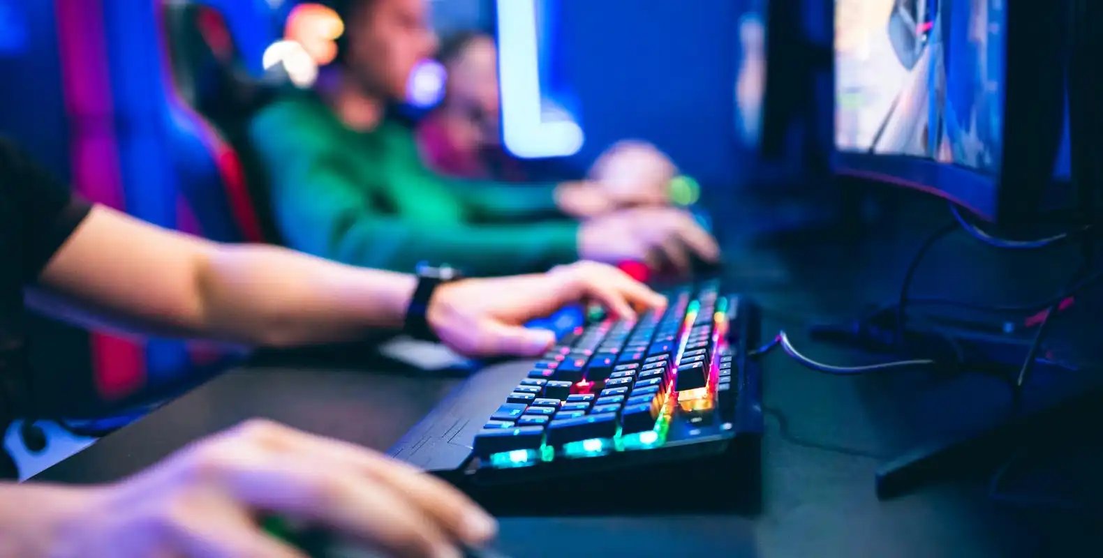 Gamers playing on computers with colorful backlit keyboards in a dimly lit room.