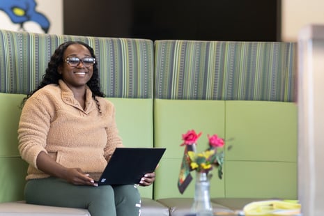 A woman wearing glasses and a tan fleece sits on a green couch, smiling while holding a laptop—perhaps researching colleges in Albany NY. In the foreground, a vase with red roses is visible on a table.