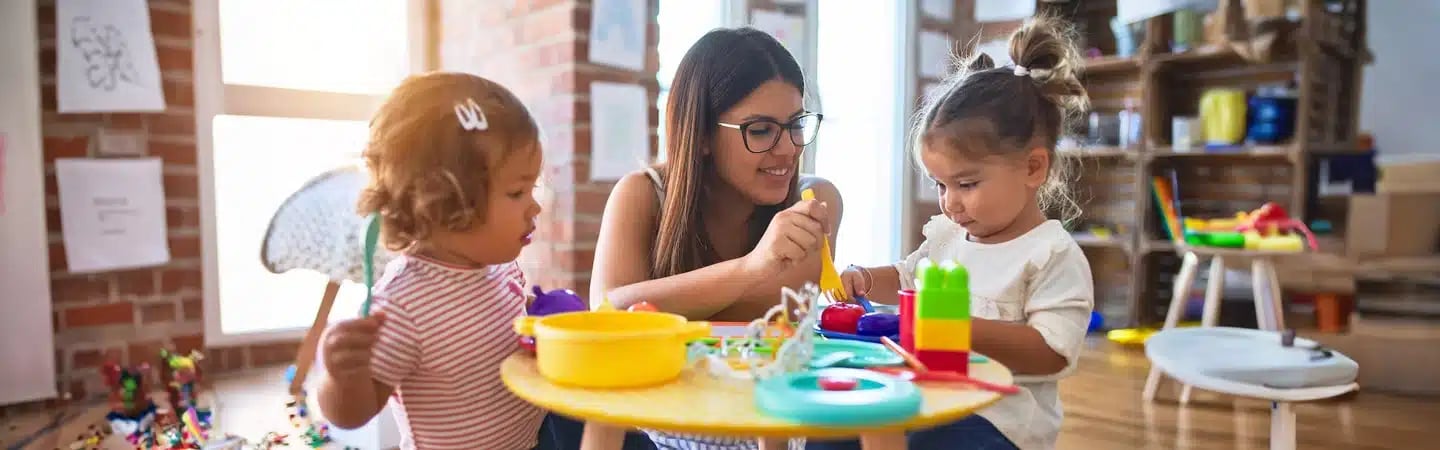 A woman plays with two young girls at a table filled with colorful toys in a bright room.