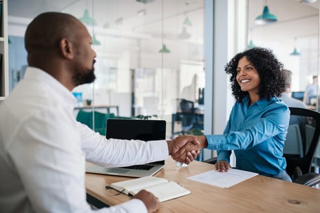 Two people sit across from each other at a desk in a modern office, smiling and shaking hands. A laptop, notebook, and paper are on the desk, suggesting a successful meeting or job interview for those with HR degrees.
