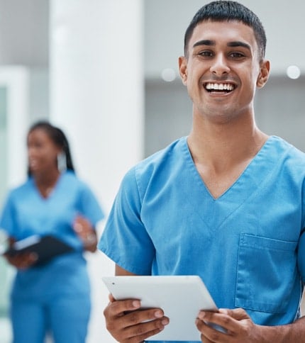 A smiling healthcare worker in blue scrubs holds a tablet, standing indoors. Another medical professional is visible in the background, also wearing blue scrubs and holding a clipboard.