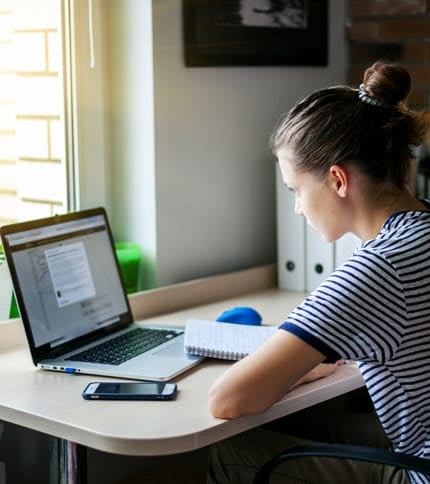 A person with hair in a bun, wearing a striped shirt, is sitting at a desk using a laptop. They are writing in a notebook, and there's a smartphone on the desk. Sunlight is streaming in through a nearby window.