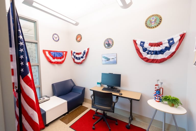 Small office decorated with patriotic bunting, military emblems on the wall, and an American flag. There’s a desk with a computer, a chair, a blue armchair, and a small table with patriotic decorations.