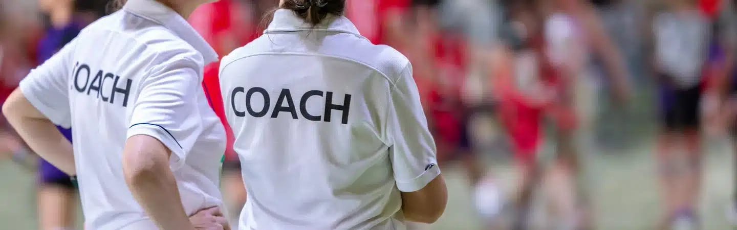 Two coaches in white shirts with "COACH" written on the back, observing a group of people in the background.