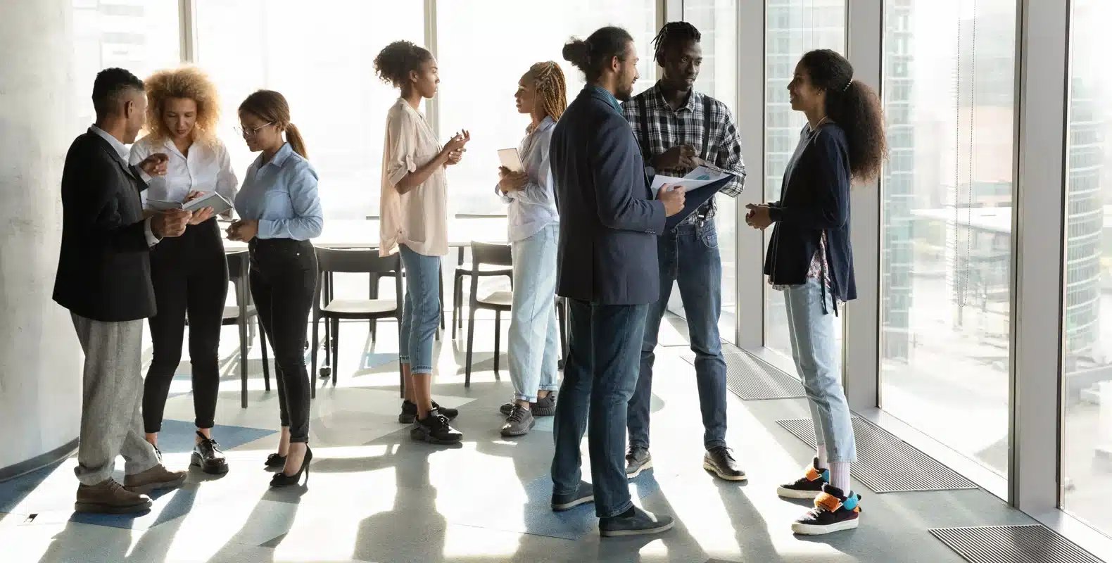 A group of people stand in pairs, chatting and holding documents in a modern office space with large windows.