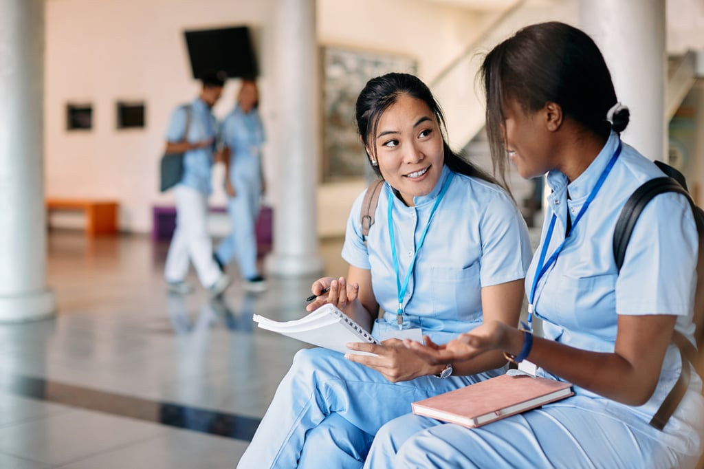 Two female students in light blue uniforms sit indoors, talking and smiling while holding notebooks about their HR degrees. In the background, other students in similar attire are walking and conversing.