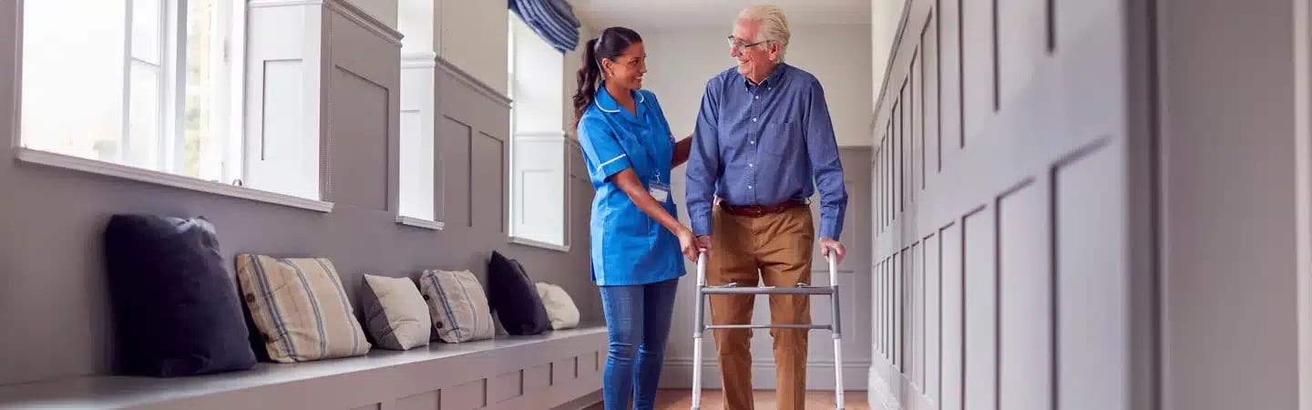 Healthcare worker assists an elderly man using a walker down a bright corridor lined with windows and cushioned seats.