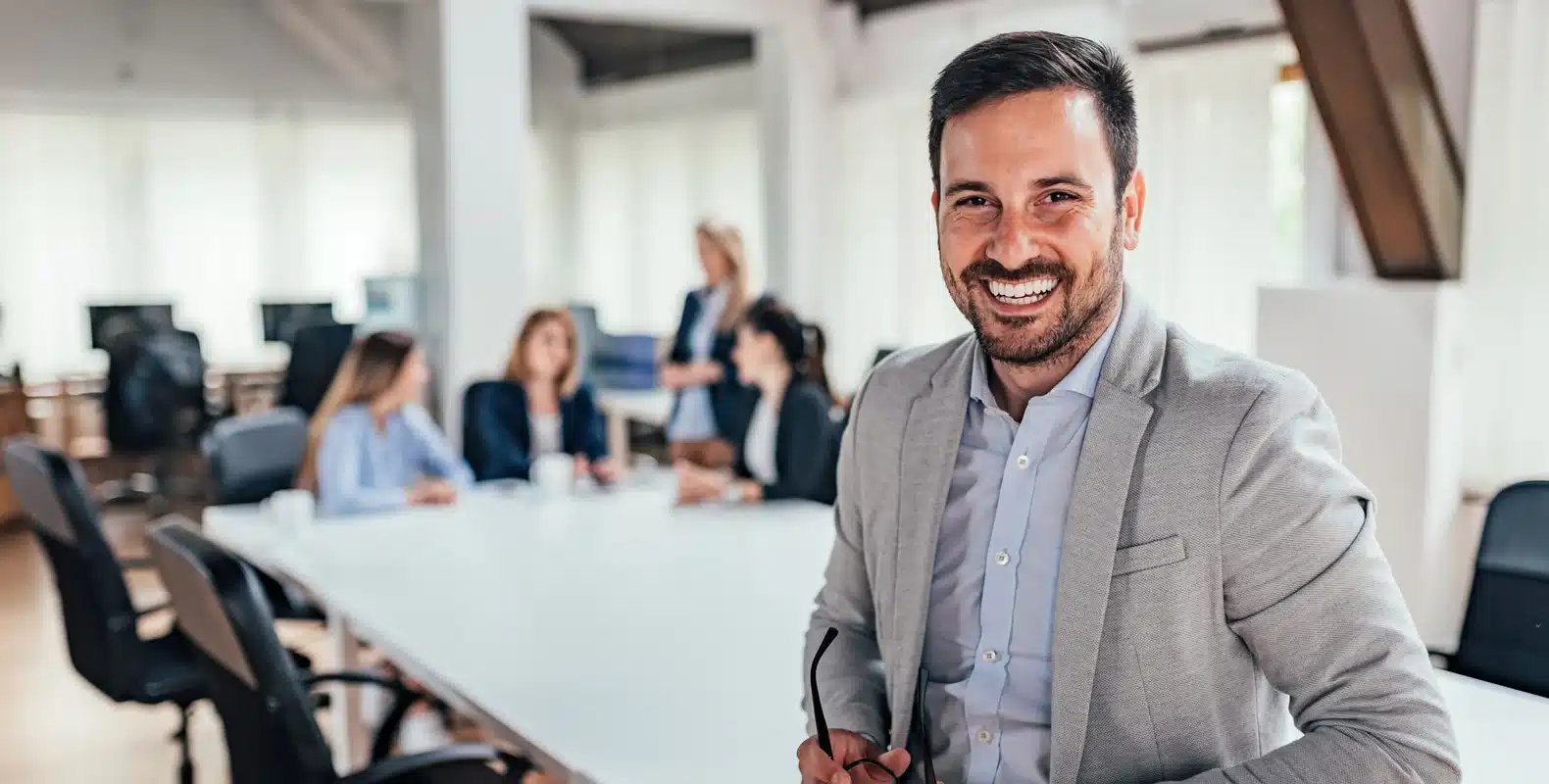 Smiling man in a gray blazer stands in an office, with colleagues gathered around a table in the background.