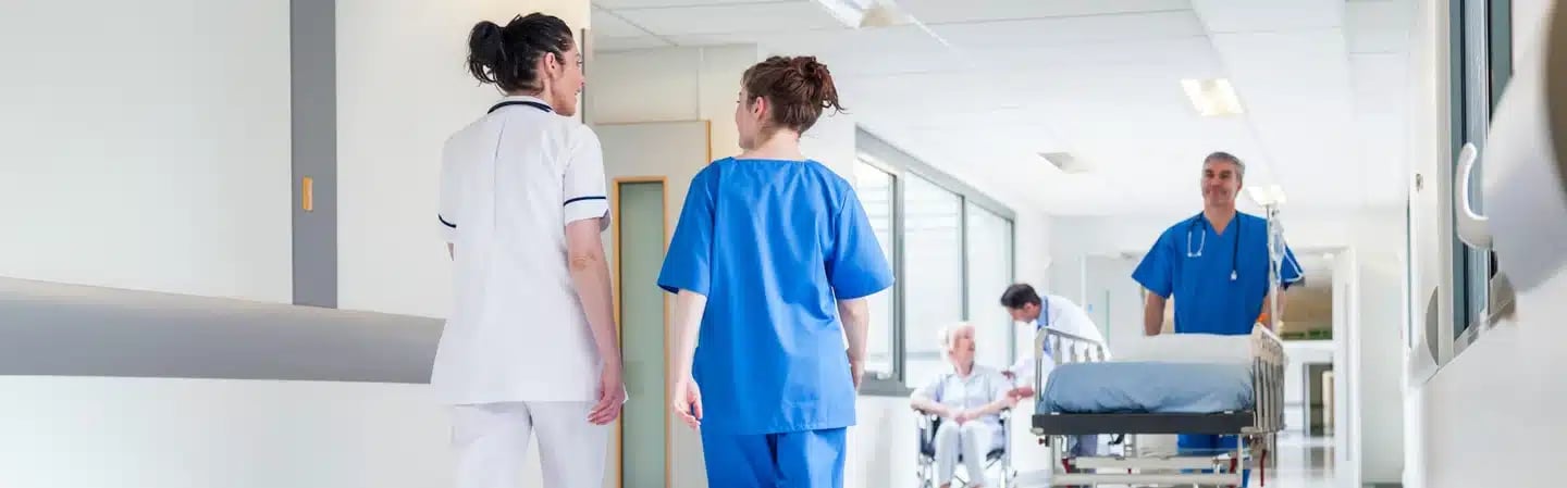 Healthcare professionals walking and conversing in a hospital hallway. In the background, a patient is being wheeled on a gurney.