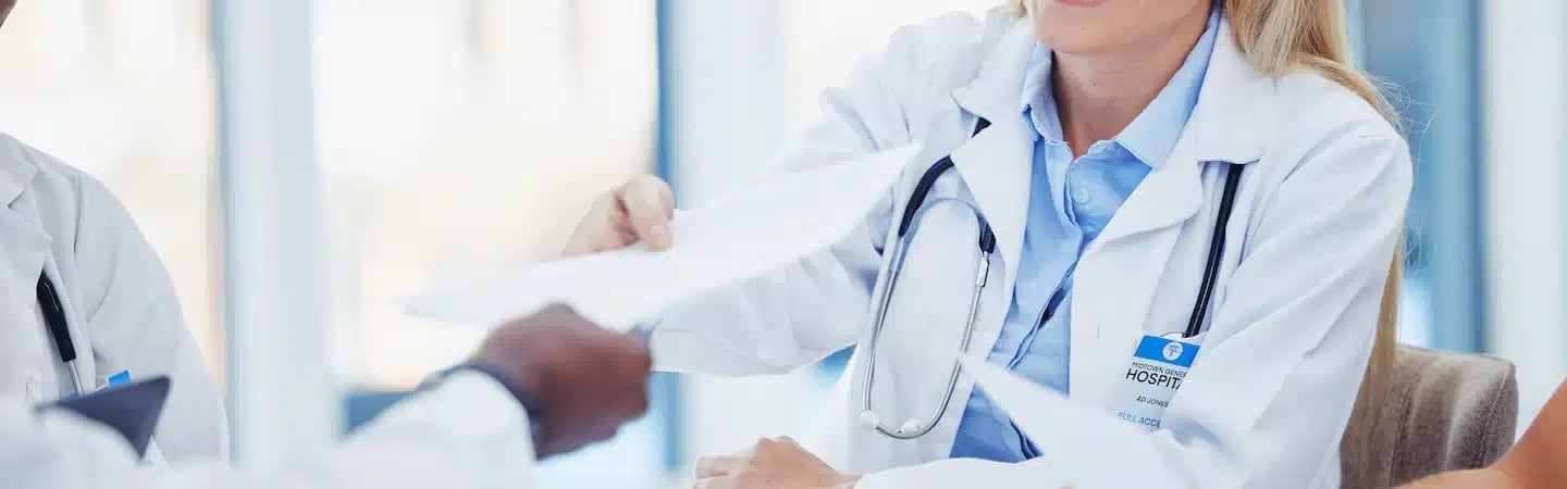 Doctors in lab coats exchanging documents in a bright room, focusing on a female doctor with a stethoscope, smiling.
