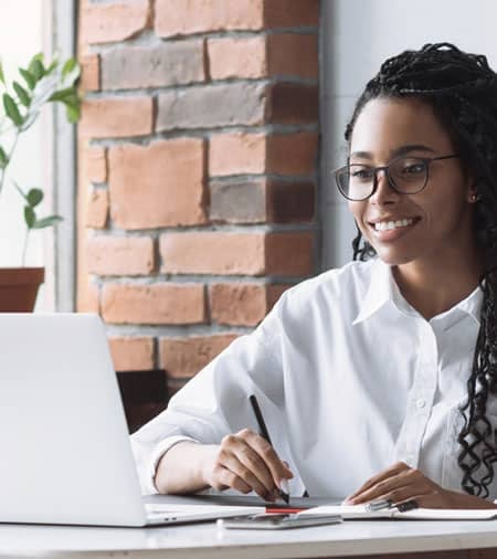A woman wearing glasses and a white shirt smiles while working at a desk with a laptop, notebook, and pen. She sits near a brick wall and a window with a potted plant visible in the background.