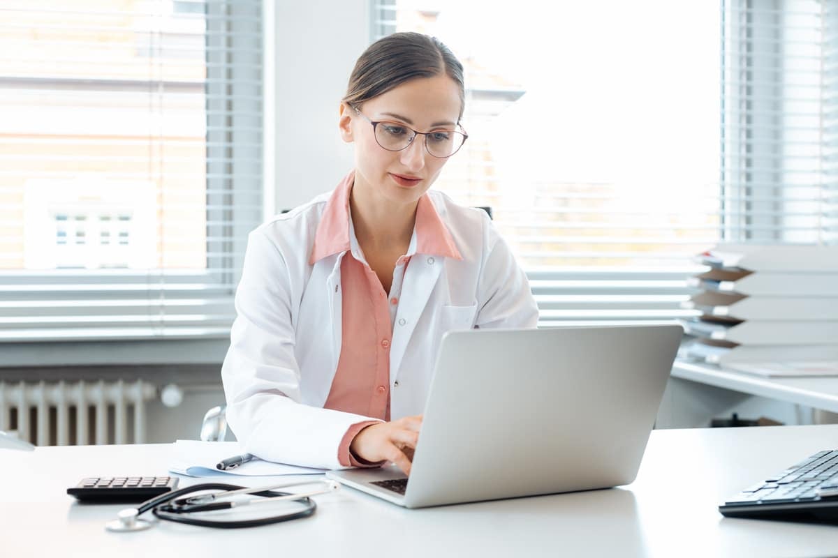 A doctor wearing glasses and a white coat is sitting at a desk, working on a laptop in a bright office.