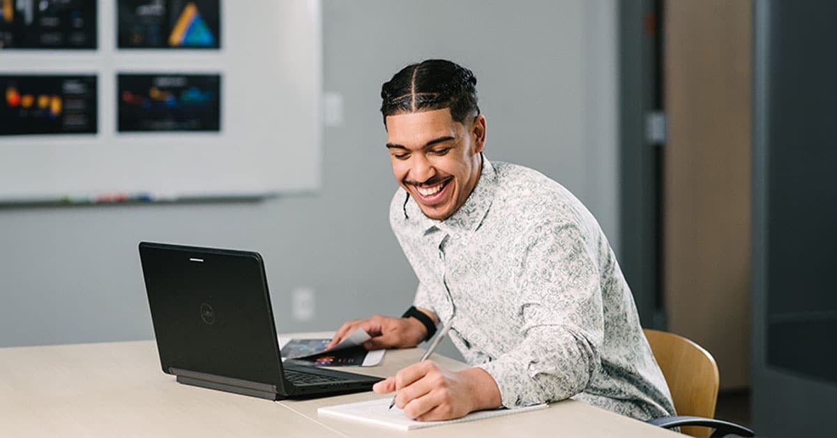 A person smiling while using a laptop and writing in a notebook at a desk in an office setting.