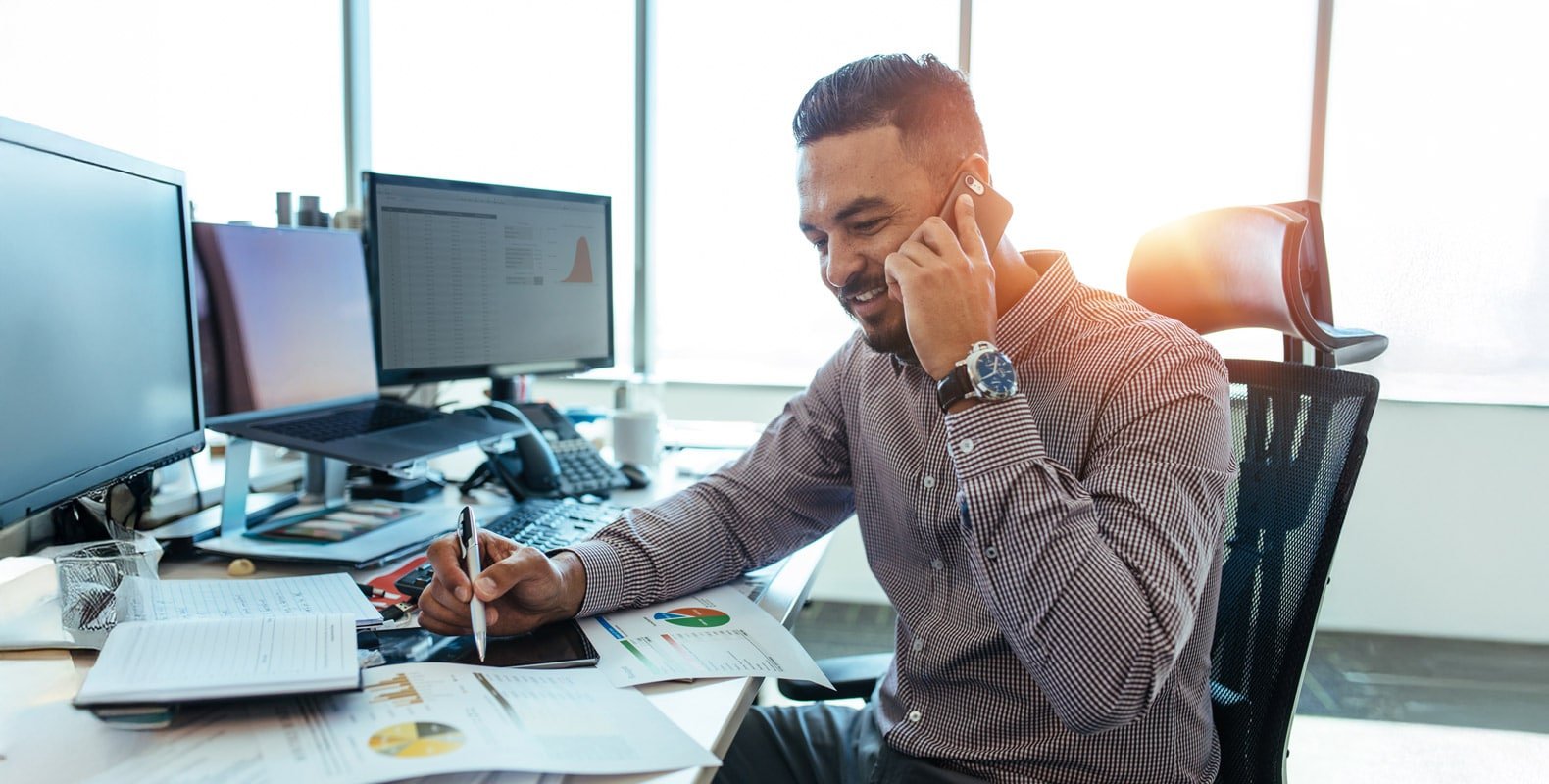 A man in a business casual shirt sits at a desk, talking on the phone while reviewing charts and notes, with multiple computer monitors in the background.