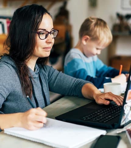A woman wearing glasses works on a laptop while taking notes in a notebook. A young boy sits beside her, focused on writing or drawing. The setting appears to be a cozy indoor space with books and a wooden table.
