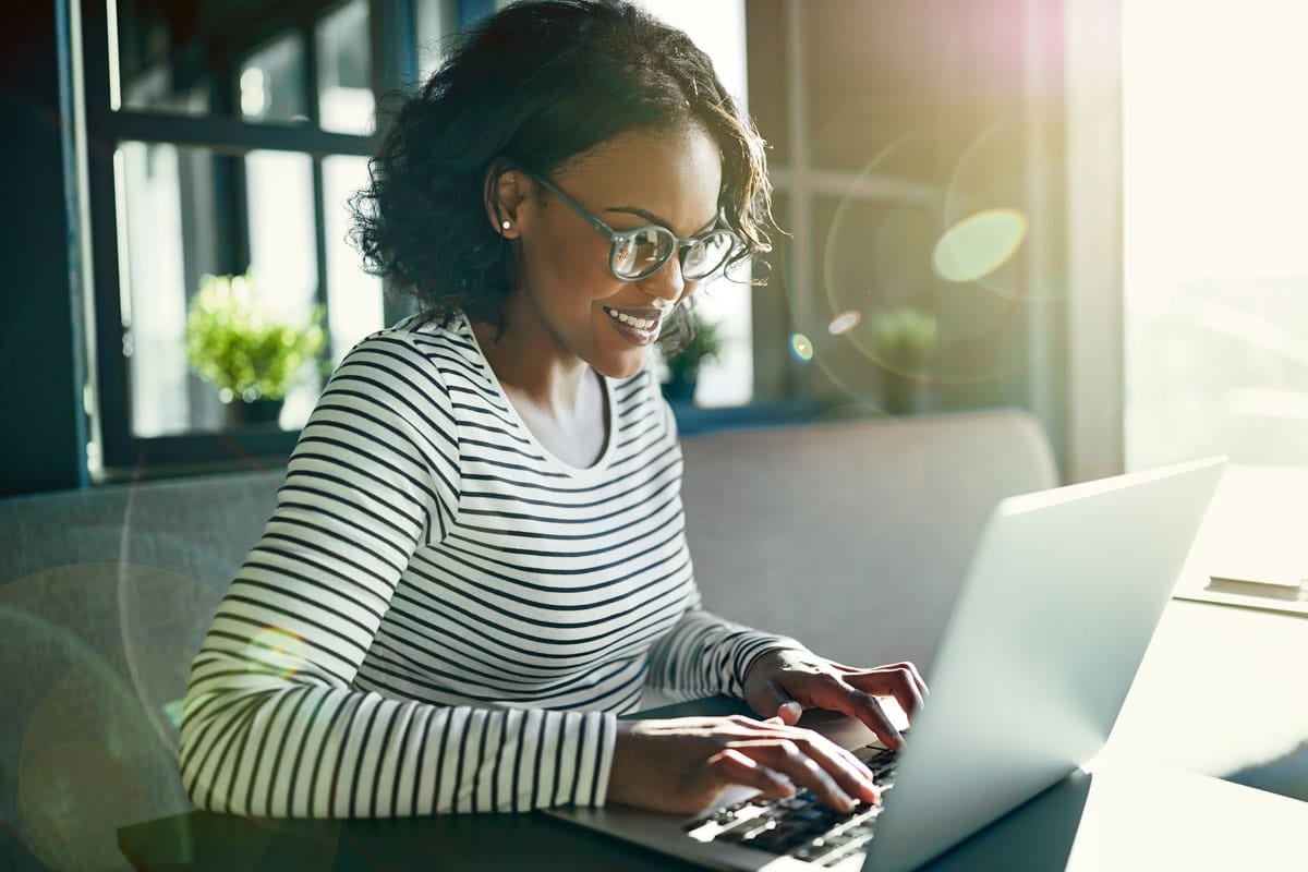A woman in a striped shirt and glasses smiles while typing on a laptop in a sunlit room.