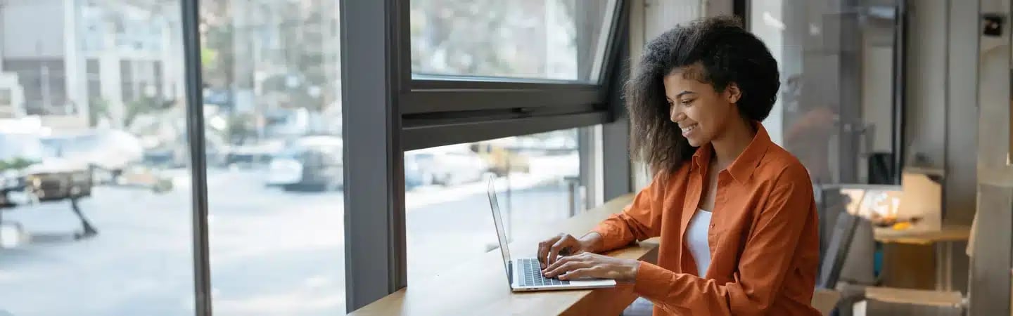 A woman is sitting at a counter near large windows, typing on a laptop with a smile.