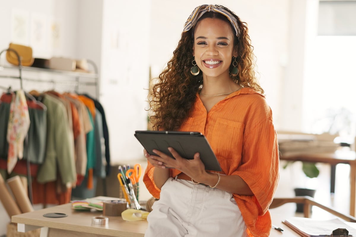 A smiling woman wearing an orange shirt holds a tablet while standing in a bright workspace, with clothing in the background.