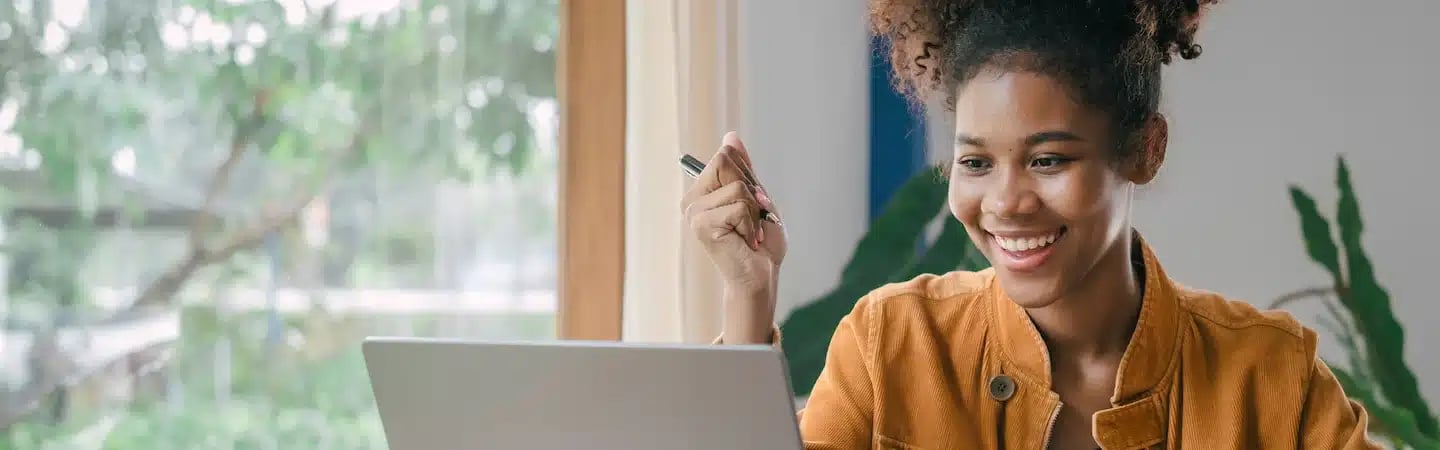 Smiling woman in a mustard jacket holding a pen and looking at her laptop in a bright, nature-filled room.
